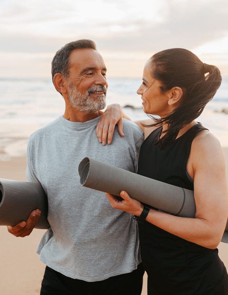 Middle-aged couple holding a yoga mat by the beach.