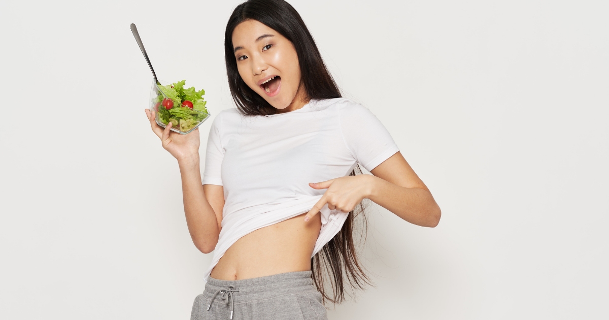 A cheerful woman in casual clothes holding a bowl of salad and pointing to her flat stomach, smiling happily against a plain white background.