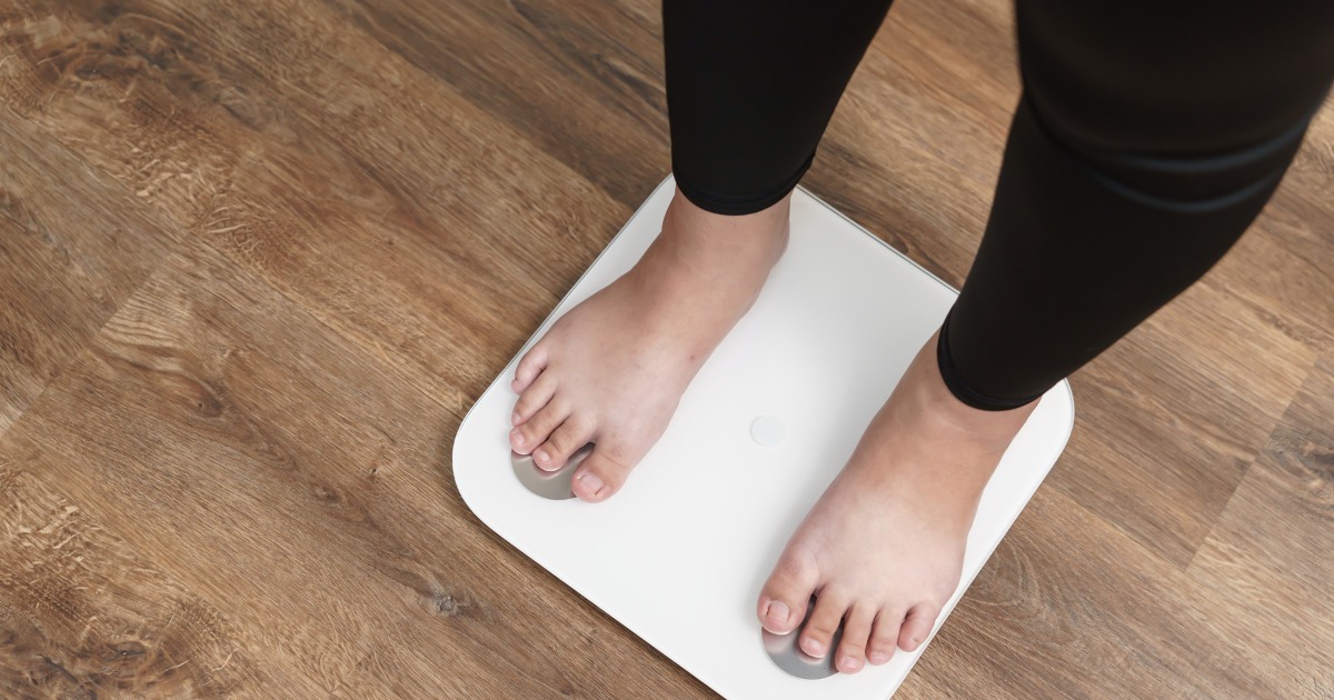 A person standing on a white digital weighing scale with bare feet, wearing black leggings, on a wooden floor.