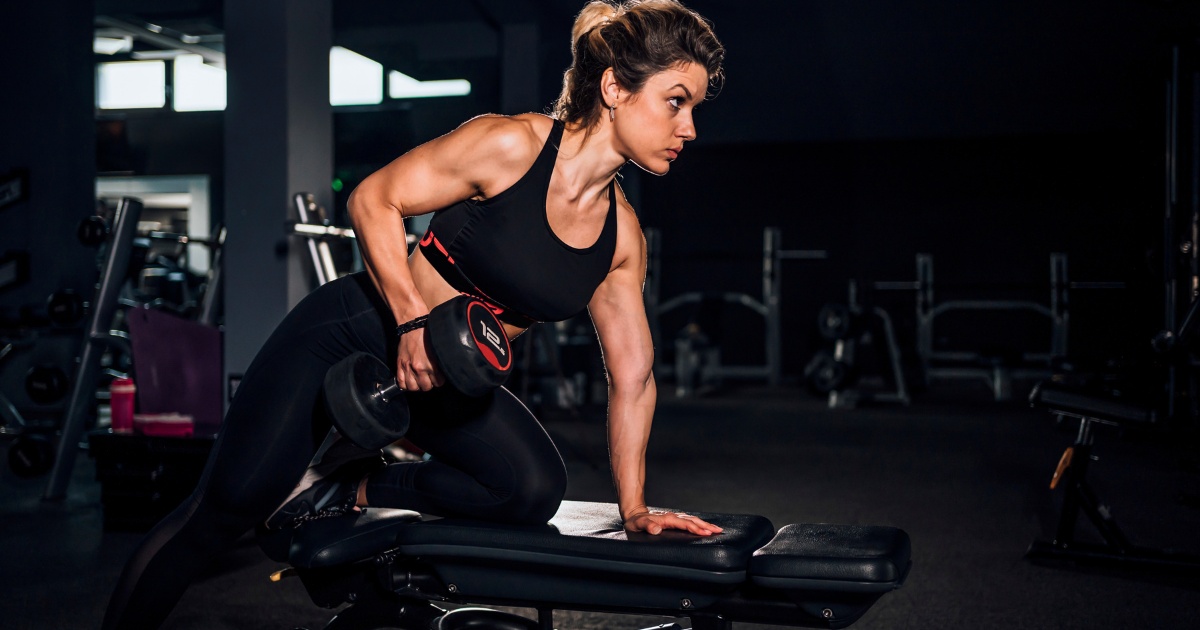 A woman in athletic wear performing a one-arm dumbbell row exercise on a gym bench, focused and lifting a weight in a dimly lit fitness center.