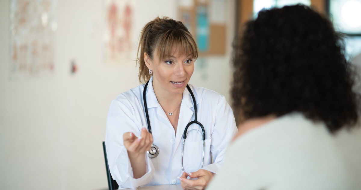 A female doctor wearing a white coat and stethoscope is talking to a patient, gesturing with her hand while listening attentively in a medical office setting.