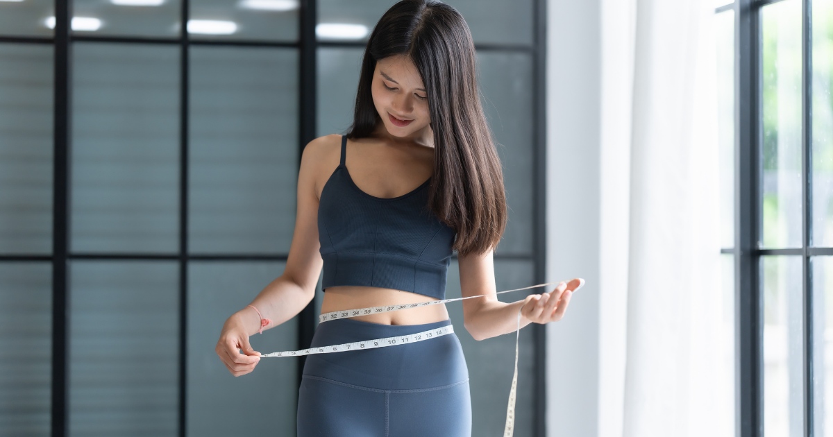 A woman in athletic wear stands indoors while measuring her waist with a tape measure, smiling as she looks down at the measurement.